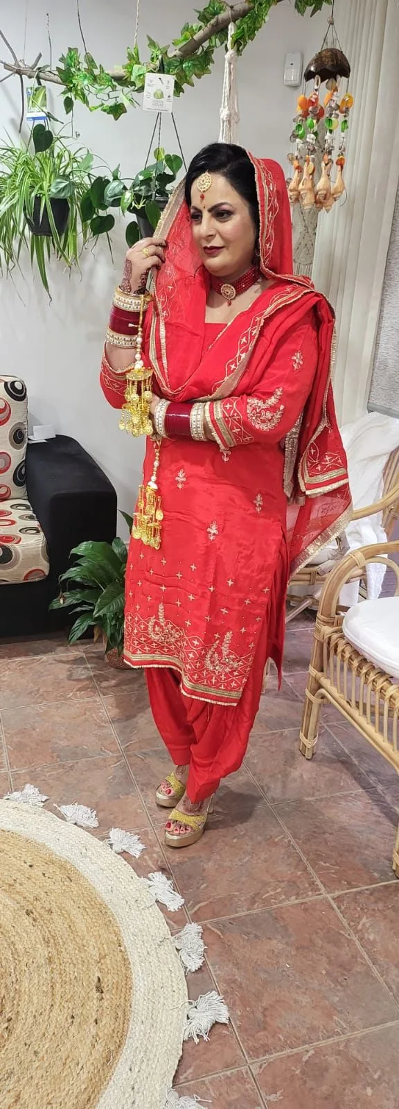 Full-length red Punjabi bridal styling with kaleere, jewellery and mehndi, inside the salon — plants, macramé and rattan chair visible